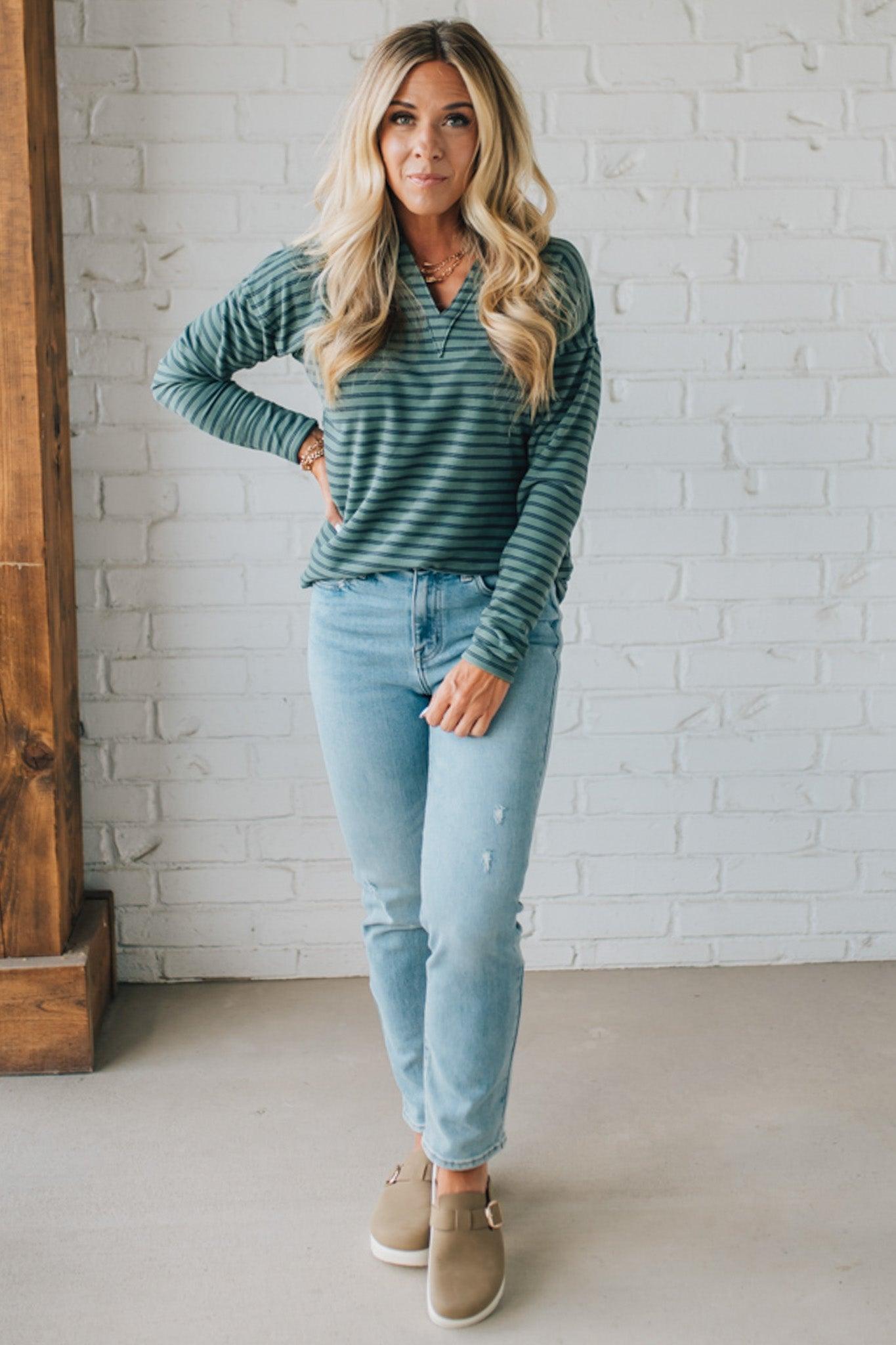 Woman wearing a green striped shirt and light blue jeans against a white brick wall.
