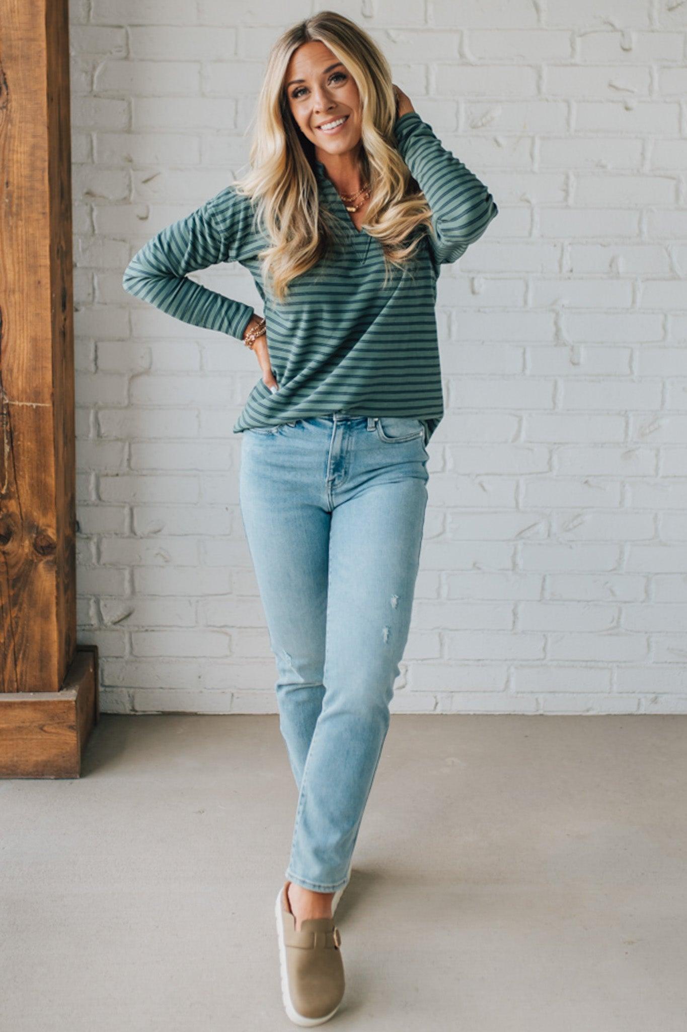 Woman wearing a striped long-sleeve top and light blue jeans standing against a white brick wall.