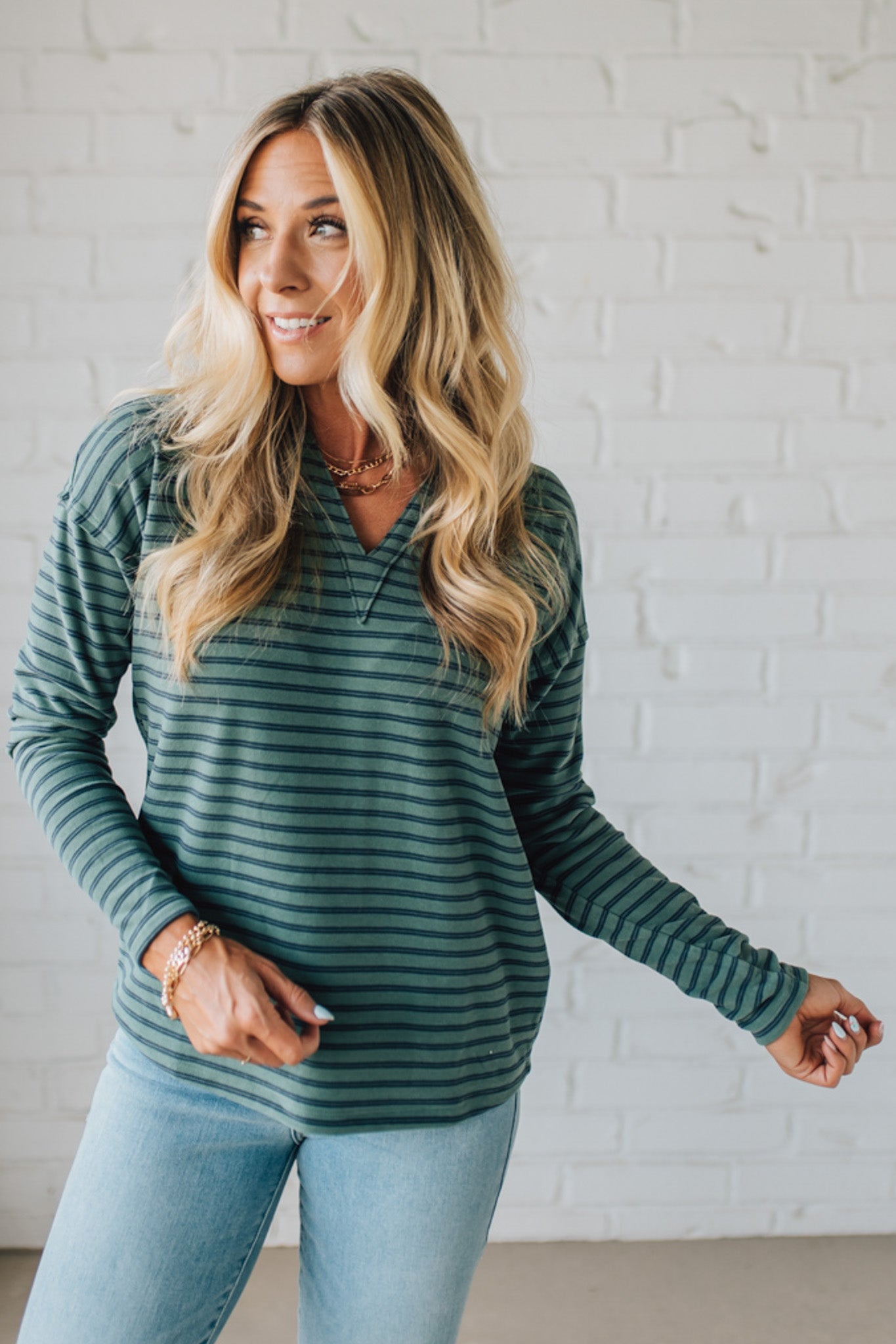 Woman wearing a green striped long-sleeve shirt and light blue jeans against a white brick wall.