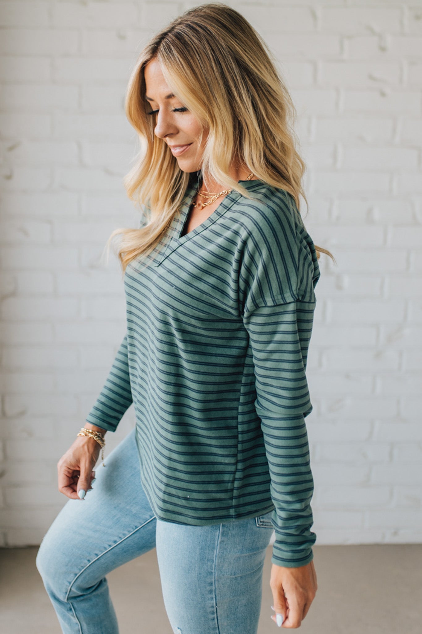 Woman wearing a green striped long-sleeve top and blue jeans against a white brick wall.