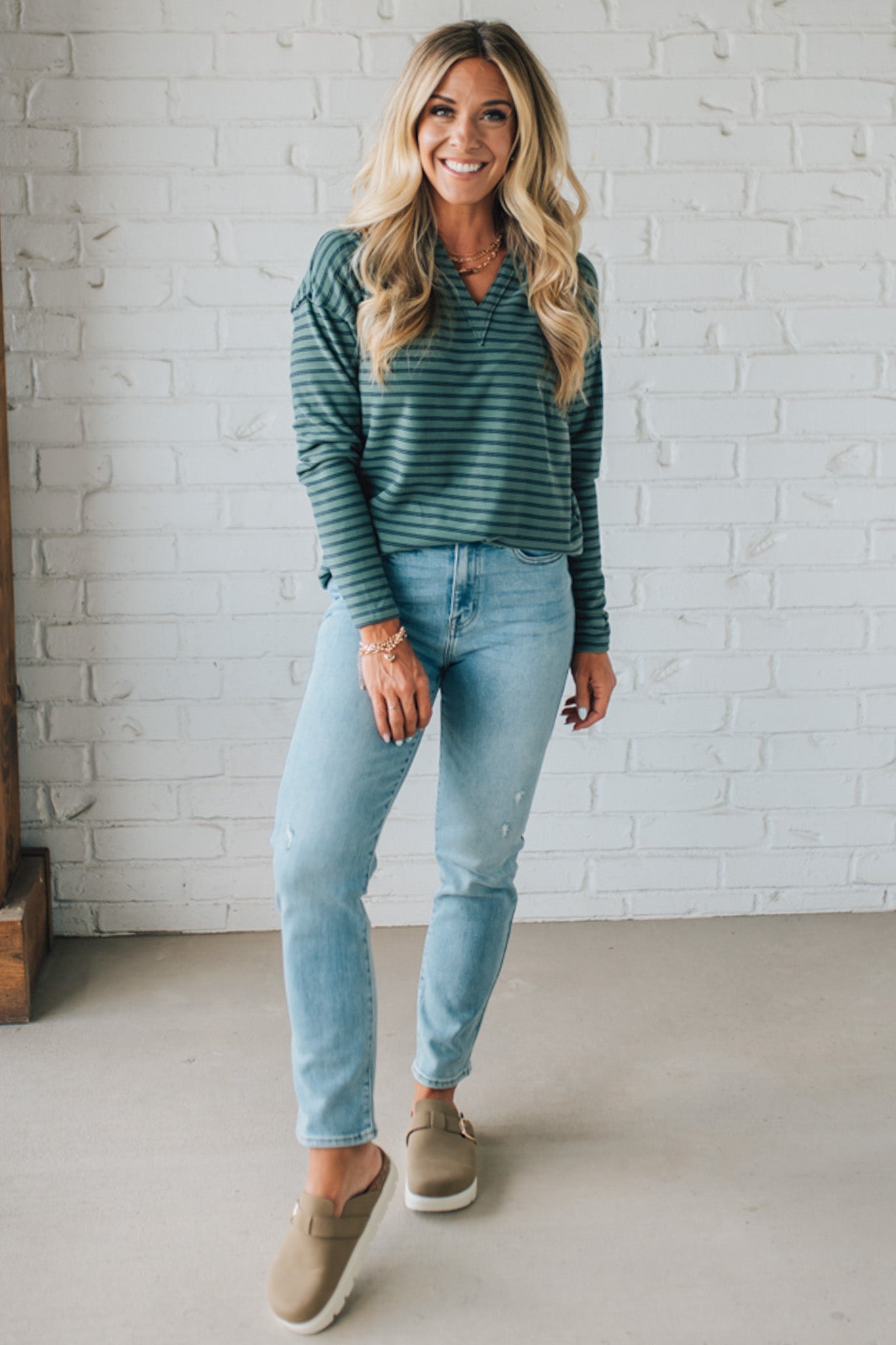 Woman wearing a green striped shirt and light blue jeans standing against a white brick wall.