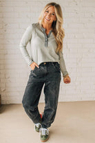 Woman wearing a gray long-sleeve shirt and black jeans against a white brick wall.