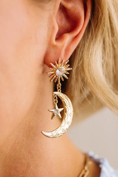 Gold earrings with sun, moon, and star designs worn by a person.