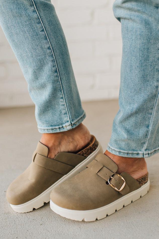 Brown clogs with a buckle worn with light blue jeans on a neutral background