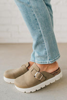 Person wearing beige clogs with white soles and light blue jeans on a neutral background