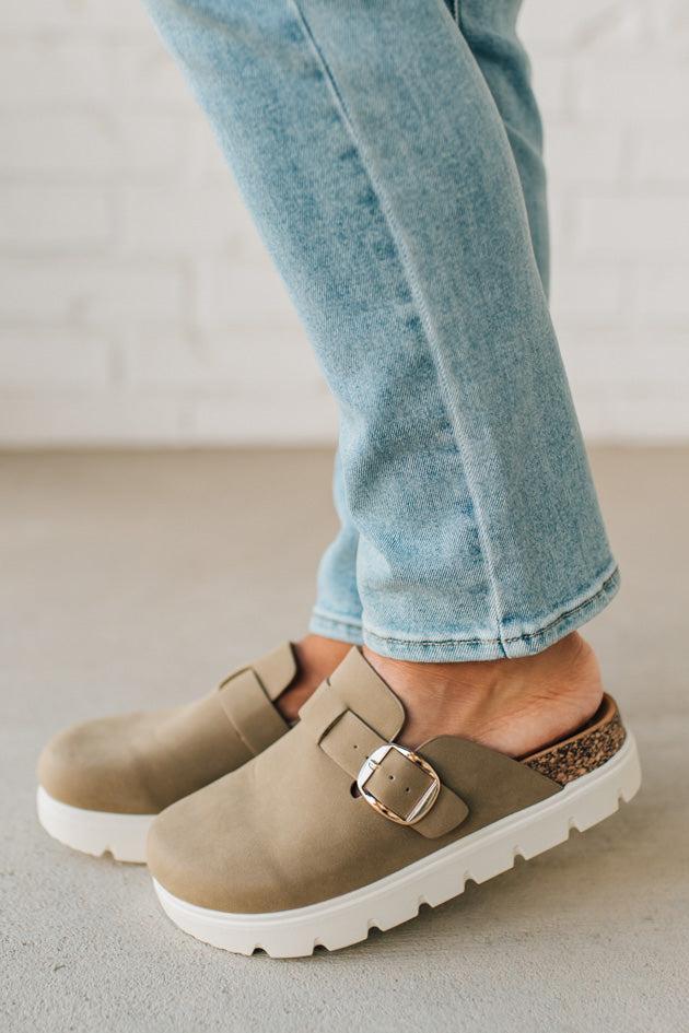 Person wearing beige clogs with white soles and light blue jeans on a neutral background