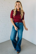 Woman wearing a red top and blue jeans standing against a white brick wall.