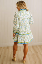 Woman wearing a floral dress standing against a light wooden paneled wall.