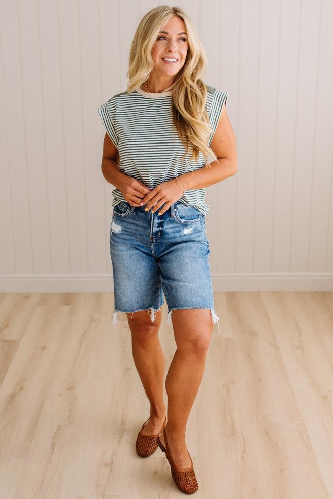 Woman wearing a striped shirt and denim shorts standing against a light wooden paneled wall.