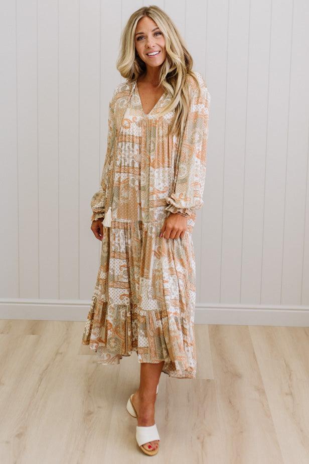 Woman wearing a long, patterned dress standing against a white wall.