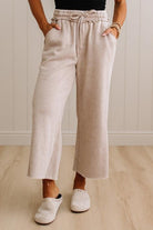 A person wearing light beige wide-leg cropped pants with an elastic drawstring waist, standing on a light wood floor against a white paneled wall.
