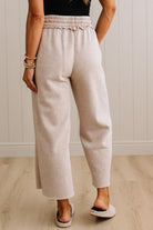 A person wearing light beige wide-leg cropped pants with an elastic waistband, shown from the back against a white paneled wall and light wood floor.