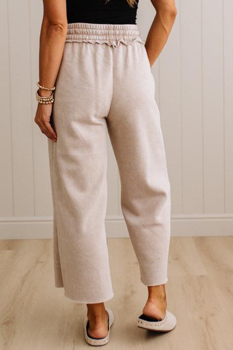 A person wearing light beige wide-leg cropped pants with an elastic waistband, shown from the back against a white paneled wall and light wood floor.