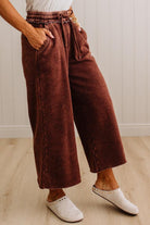A person wearing wide-leg cropped pants in a dark rust brown corduroy-like fabric, paired with white slip-on shoes, standing on a light wood floor against a white paneled wall.