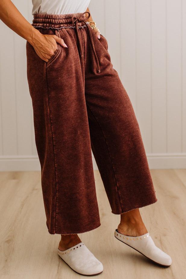 A person wearing wide-leg cropped pants in a dark rust brown corduroy-like fabric, paired with white slip-on shoes, standing on a light wood floor against a white paneled wall.