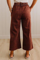 A person wearing rust-brown wide-leg cropped pants with an elastic waistband, standing on a light wood floor against a white paneled wall.