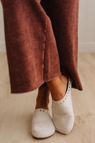 A close-up of a person's feet wearing off-white slip-on shoes with gold-tone stud detailing, paired with rust-brown wide-leg pants, standing on a light wood floor.