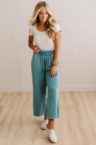 A woman modeling a pair of wide-leg cropped pants in a dusty teal color, paired with a white t-shirt and white slip-on shoes, standing in a room with light wood floors and white paneled walls.