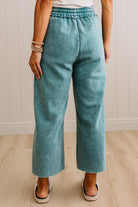 A person wearing high-waisted, wide-leg cropped pants in a dusty teal or blue-green color, paired with a white top and white slide sandals, standing against a light wood-paneled wall.