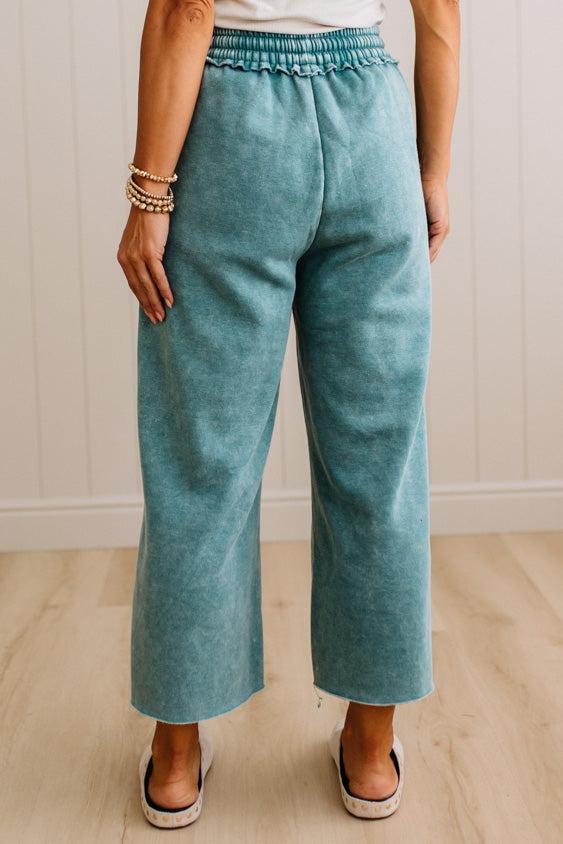 A person wearing high-waisted, wide-leg cropped pants in a dusty teal or blue-green color, paired with a white top and white slide sandals, standing against a light wood-paneled wall.
