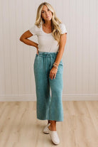 A woman modeling a white short-sleeve crewneck t-shirt and high-waisted, wide-leg teal pants with white slip-on shoes, standing in front of a white paneled wall and light wood floor.