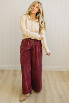 Woman wearing a beige blouse and burgundy pants standing against a white wall.