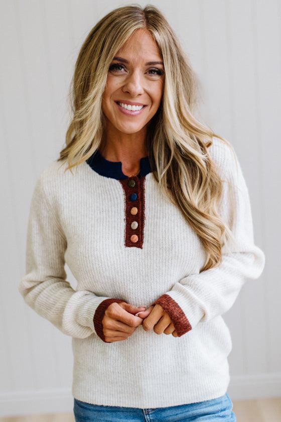 Blonde woman modeling an oat sweater with navy collar, mocha cuffs, and a mocha henlwy with multi color sweater covered buttons. Standing on a light wood floor with light backdrop.