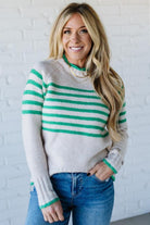 Woman wearing a gray and green striped sweater with blue jeans against a white brick wall.