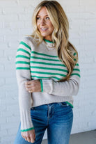 Woman wearing a gray and green striped sweater with blue jeans against a white brick wall.