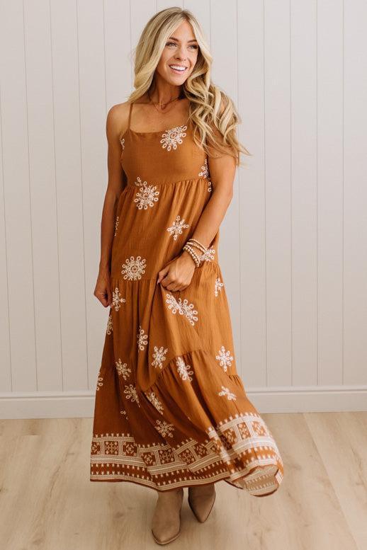 A woman modeling a rust-brown maxi dress with white floral and geometric patterns, standing against a white paneled wall and light wood floor.