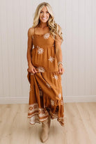 A woman modeling a rust-brown tiered maxi dress with white floral embroidery and a patterned hem, paired with beige ankle boots, set against a white paneled wall and light wood floor.