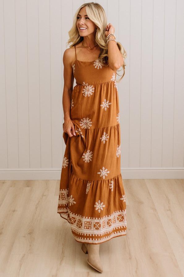 A woman modeling a rust-brown tiered maxi dress with white floral embroidery and a decorative embroidered hem, standing in a room with light wood floors and white paneled walls.