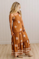 A woman modeling a rust-brown tiered maxi dress with white floral embroidery and a decorative border, paired with beige ankle boots against a white paneled wall and light wood floor.