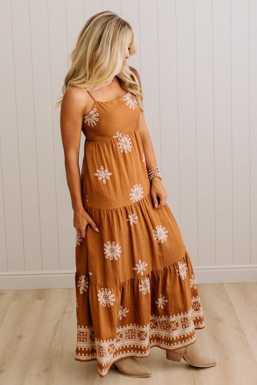A woman modeling a rust-brown tiered maxi dress with white floral embroidery and a decorative border, paired with beige ankle boots against a white paneled wall and light wood floor.