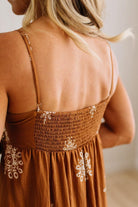 Close-up back view of a woman wearing a brown smocked dress with white embroidered details and thin straps.