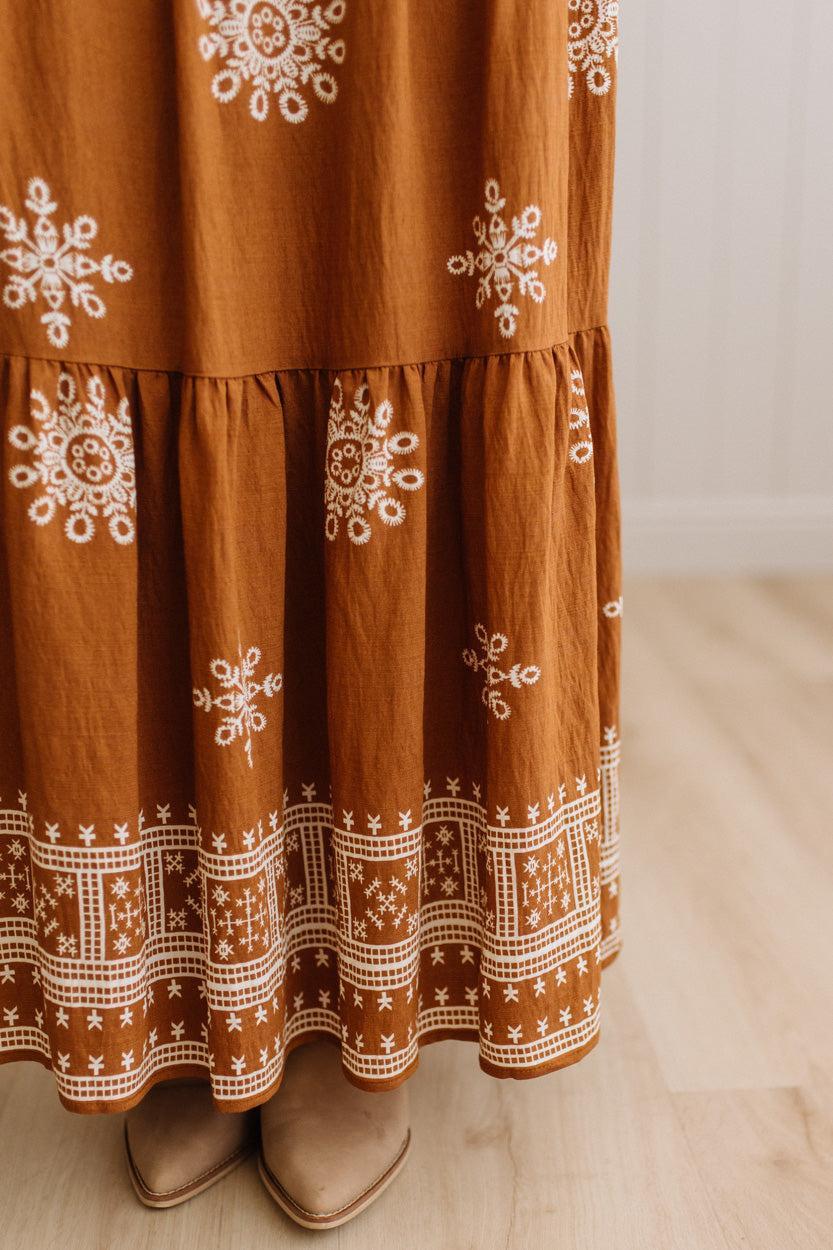 A close-up of a rust-brown tiered maxi skirt with white embroidered snowflake and geometric patterns, shown on a model wearing tan pointed-toe flats.