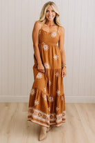 A woman modeling a rust-orange tiered maxi dress with white floral and geometric embroidery, standing against a white paneled wall and light wood floor.