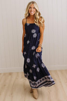 A woman modeling a navy blue maxi dress with white snowflake patterns and a decorative border at the hem, standing in a room with light wood floors and white paneled walls.