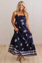 A woman modeling a navy blue maxi dress with white floral and geometric patterns, standing against a white paneled wall and light wood floor.