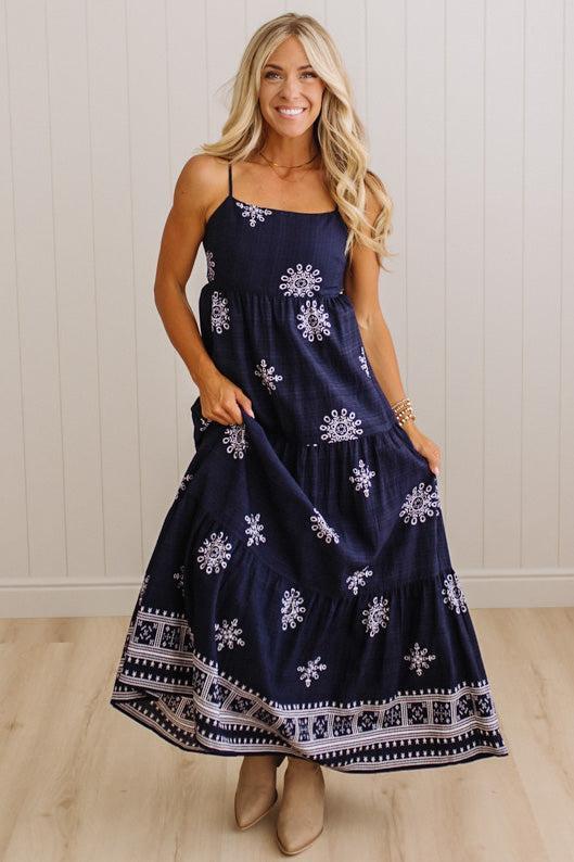 A woman modeling a navy blue maxi dress with white floral and geometric patterns, standing against a white paneled wall and light wood floor.