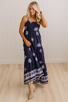 A woman modeling a navy blue maxi dress with white floral and geometric patterns, paired with beige ankle boots, standing in a room with light wood floors and white paneled walls.