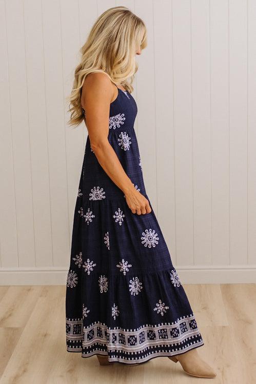 A woman wearing a sleeveless navy maxi dress with white snowflake and geometric patterns, standing in a room with light wood floors and white paneled walls.