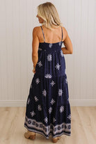 A woman wearing a navy blue maxi dress with white floral and geometric patterns, shown from the back against a white paneled wall and light wood floor.