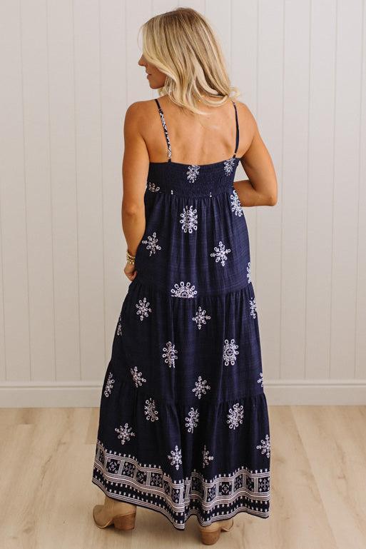 A woman wearing a navy blue maxi dress with white floral and geometric patterns, shown from the back against a white paneled wall and light wood floor.