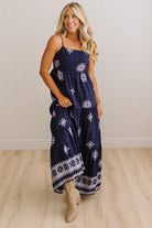 A woman modeling a navy blue maxi dress with white snowflake and geometric patterns, standing in front of a white paneled wall and light wood floor.