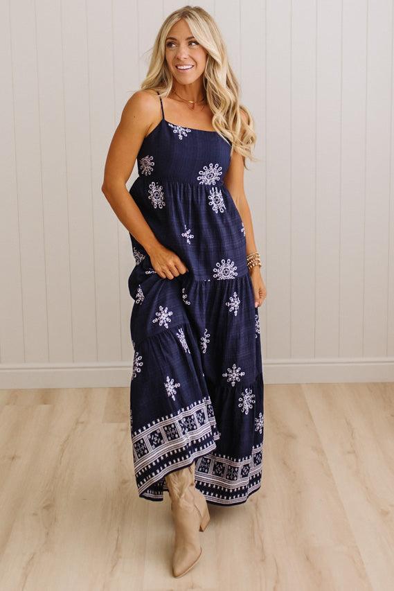 A woman modeling a navy blue maxi dress with white snowflake and geometric patterns, standing in front of a white paneled wall and light wood floor.