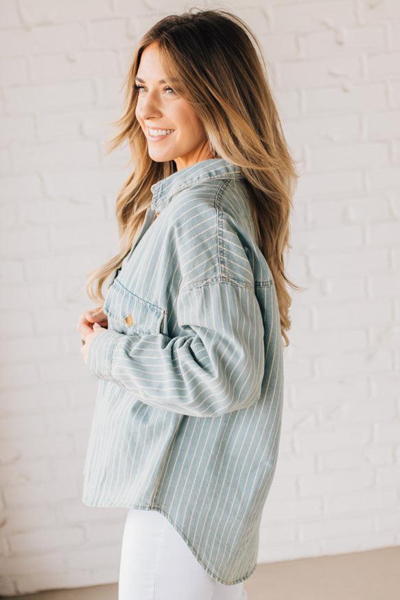 Blonde wearing a light blue denim oversized shirt with vertical white stripes and brown large buttons.