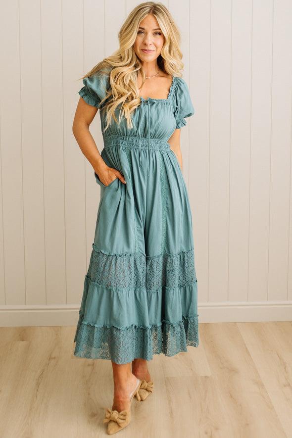 Woman wearing a teal dress with lace details in a room with wooden flooring and white paneled walls.