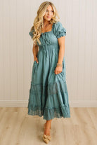 Woman wearing a teal dress with lace details against a white paneled wall.