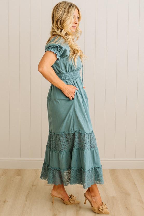 Woman wearing a teal dress with lace details against a white paneled wall.